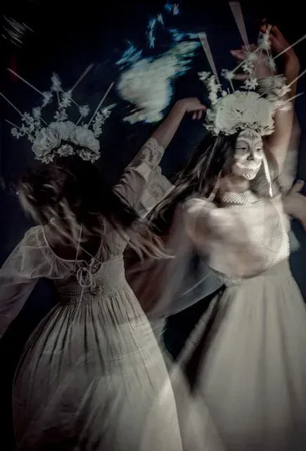 Two women in white dresses with flower crowns dancing. Dark, abstract background, ethereal.