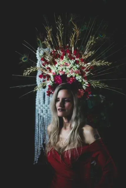 Woman in red dress with floral headdress, standing in front of a dark background.