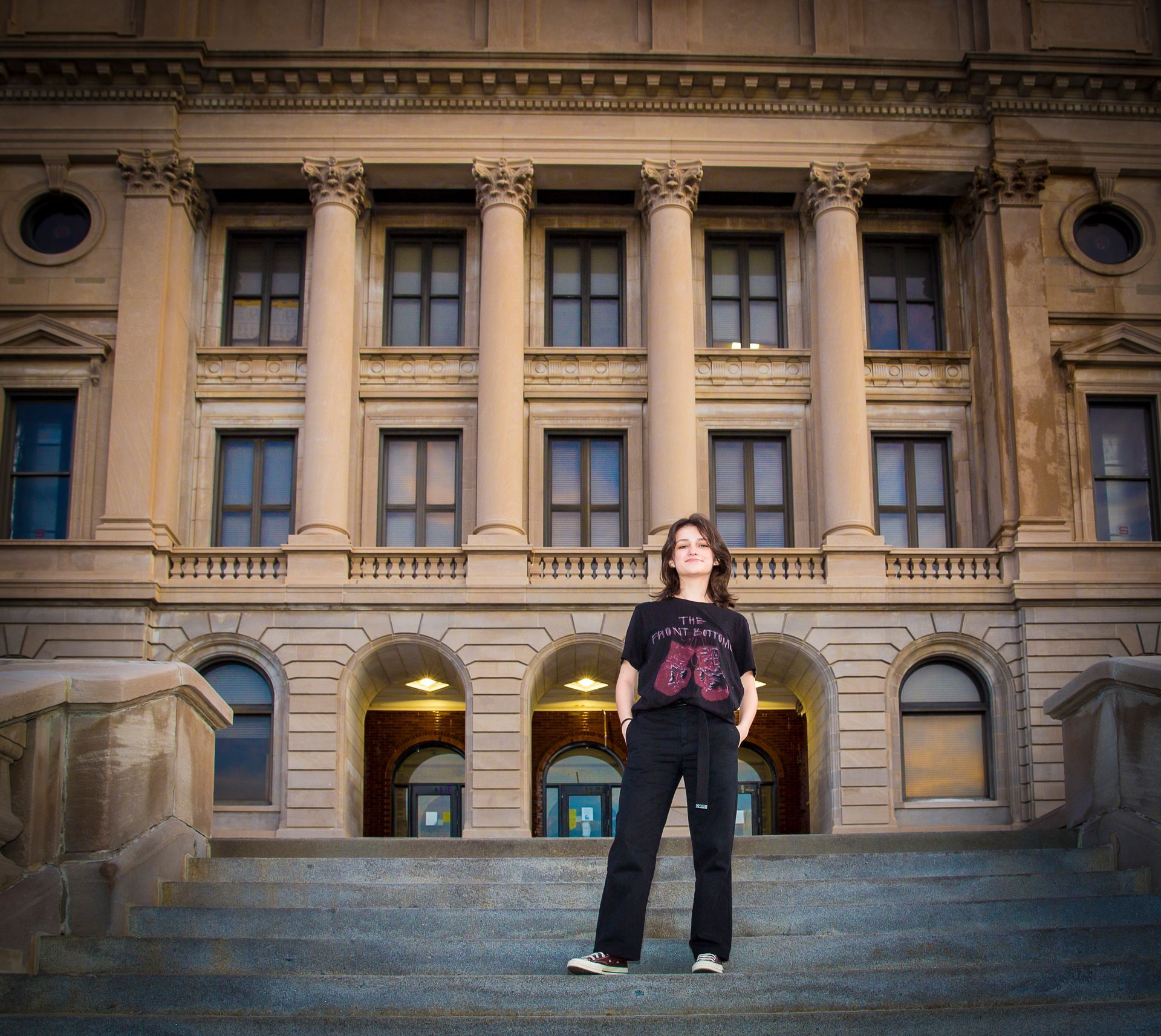 Teen stands on steps of a stately building. Wearing black pants and a t-shirt, arms at sides. Building has pillars and windows.