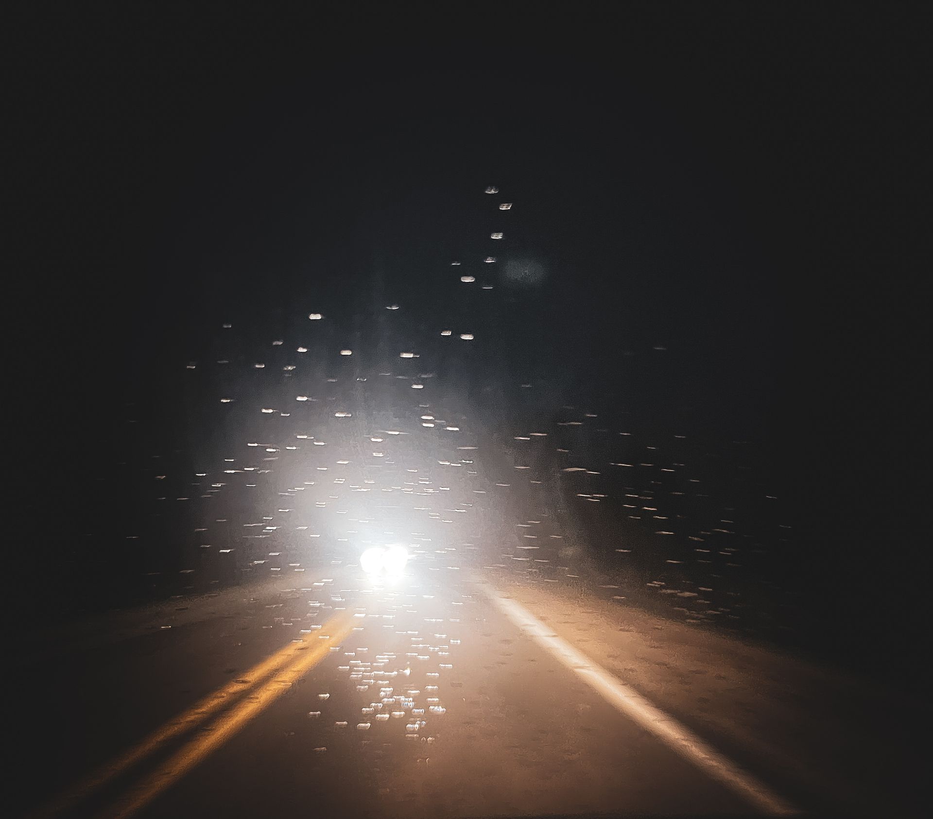 Car headlights illuminate a dark, wet road at night; water droplets on the windshield create a hazy effect.