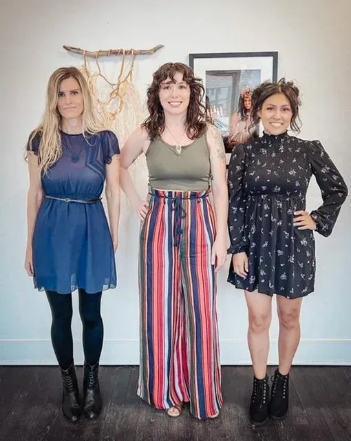 Three women standing in front of a white wall. The woman on the left wears a blue dress, the middle one wears a green top and striped pants, and the right wears a black dress.