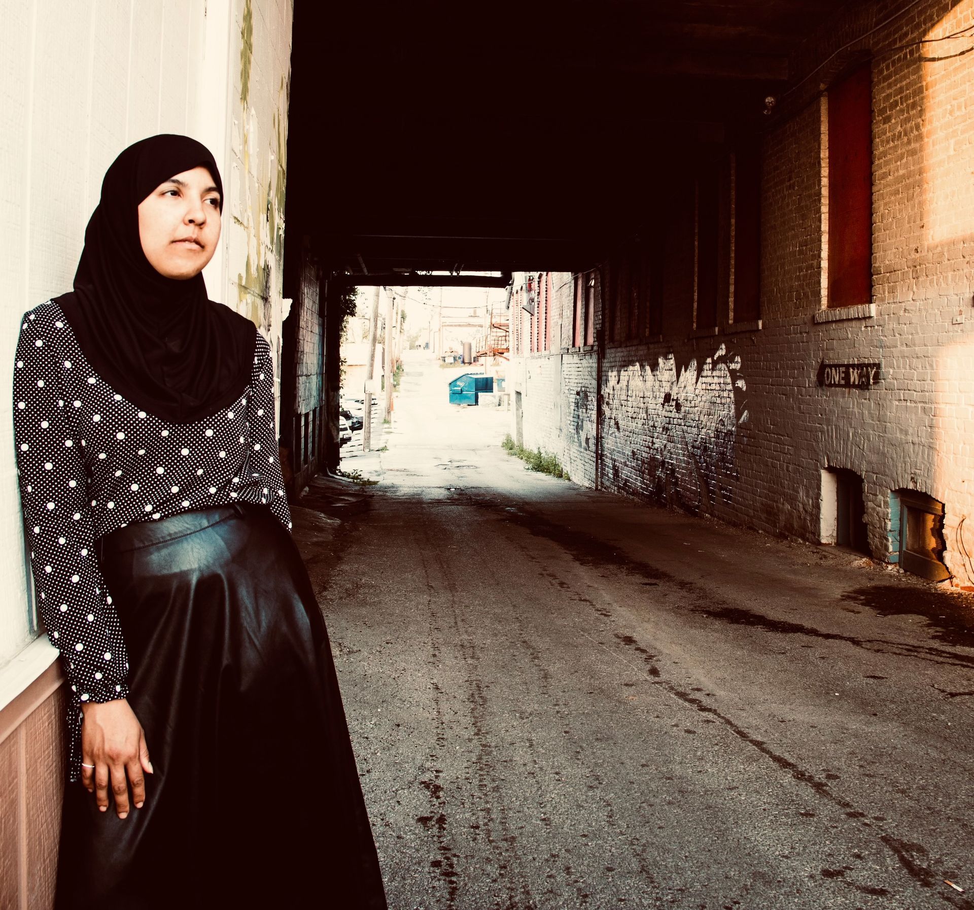 Woman in hijab leans against a wall in a dark alley; looking away.