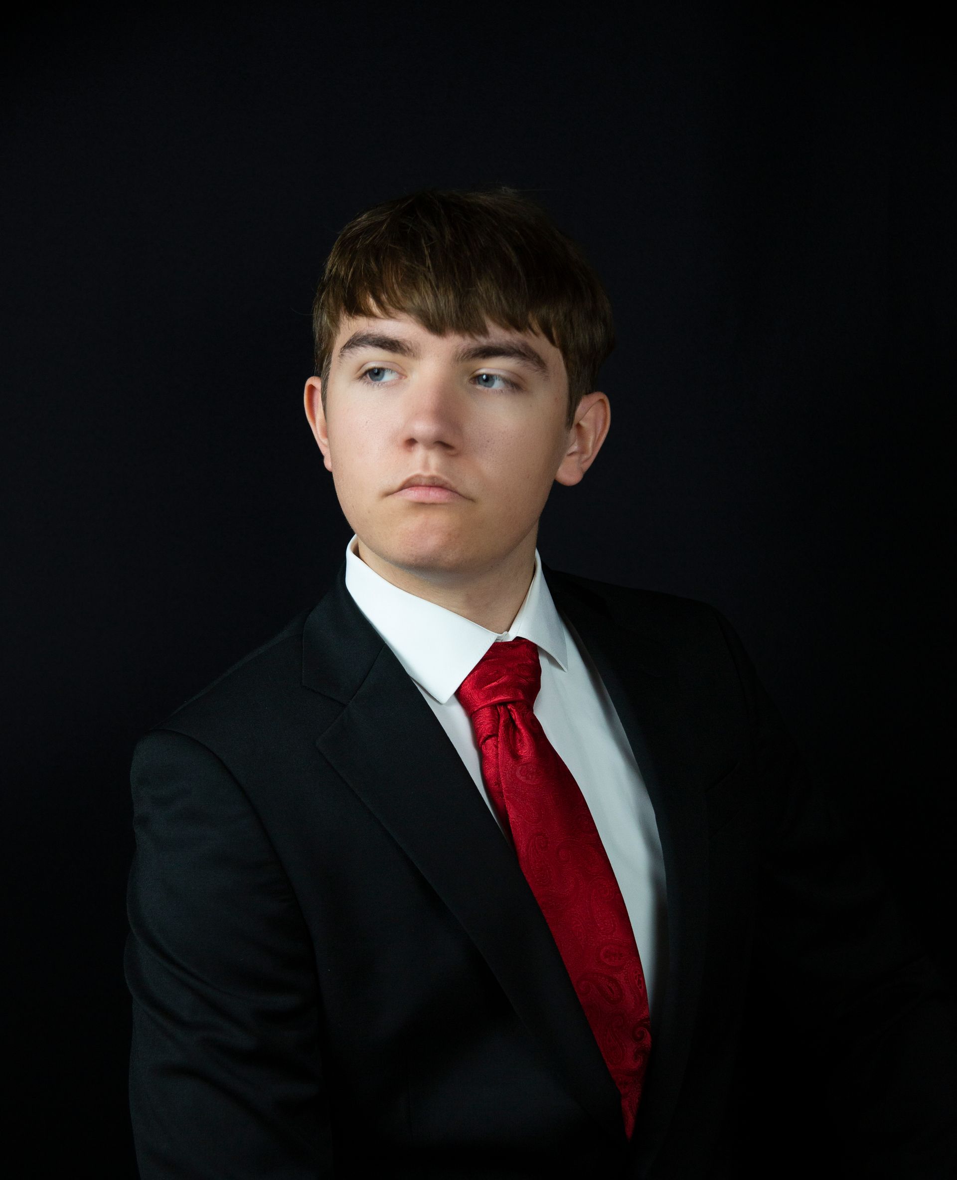 Young man in suit, red tie, looking to the side, against a black background.
