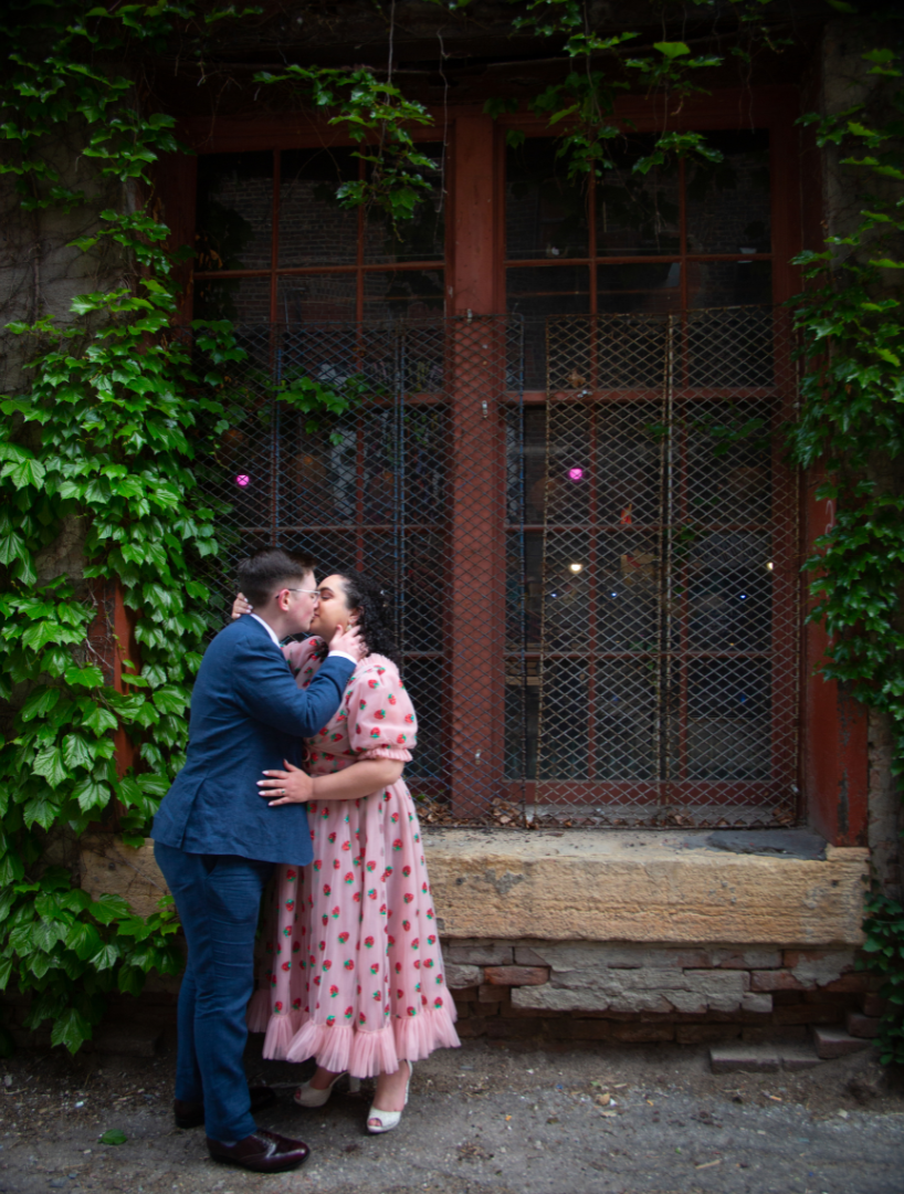 Couple kissing in front of a brick building with a window covered in vines. The woman wears a pink dress, the man a blue suit.