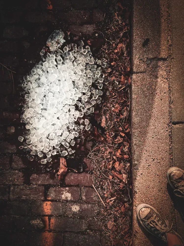 Ice cubes scattered on brick and concrete, near a sidewalk; person's feet in sneakers are visible.