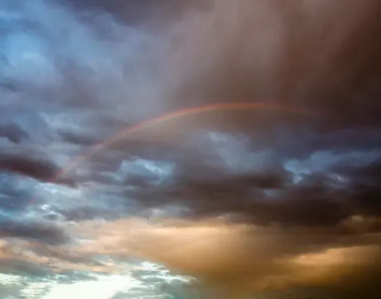 Rainbow arcs across a cloudy sky, with hues of orange, blue, and gray.