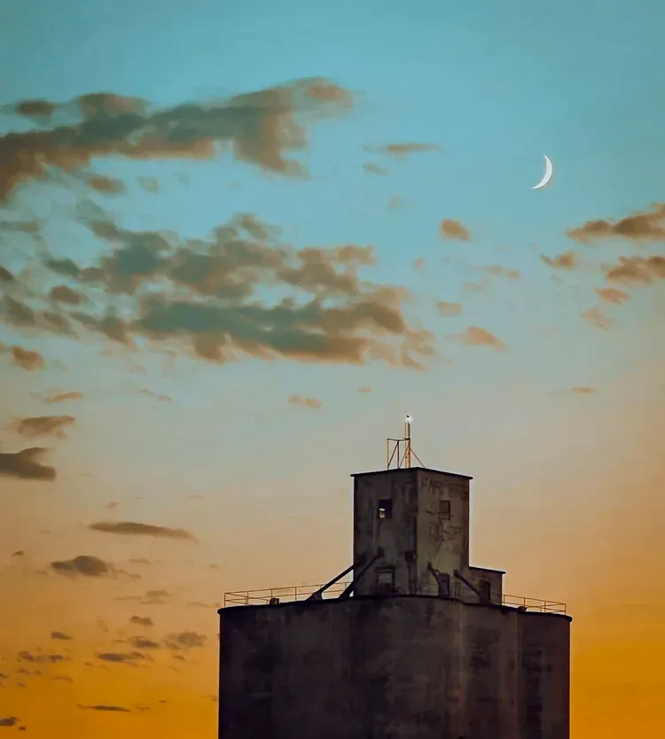 Old grain silo against a sky of orange, blue, and clouds; a crescent moon hangs above.