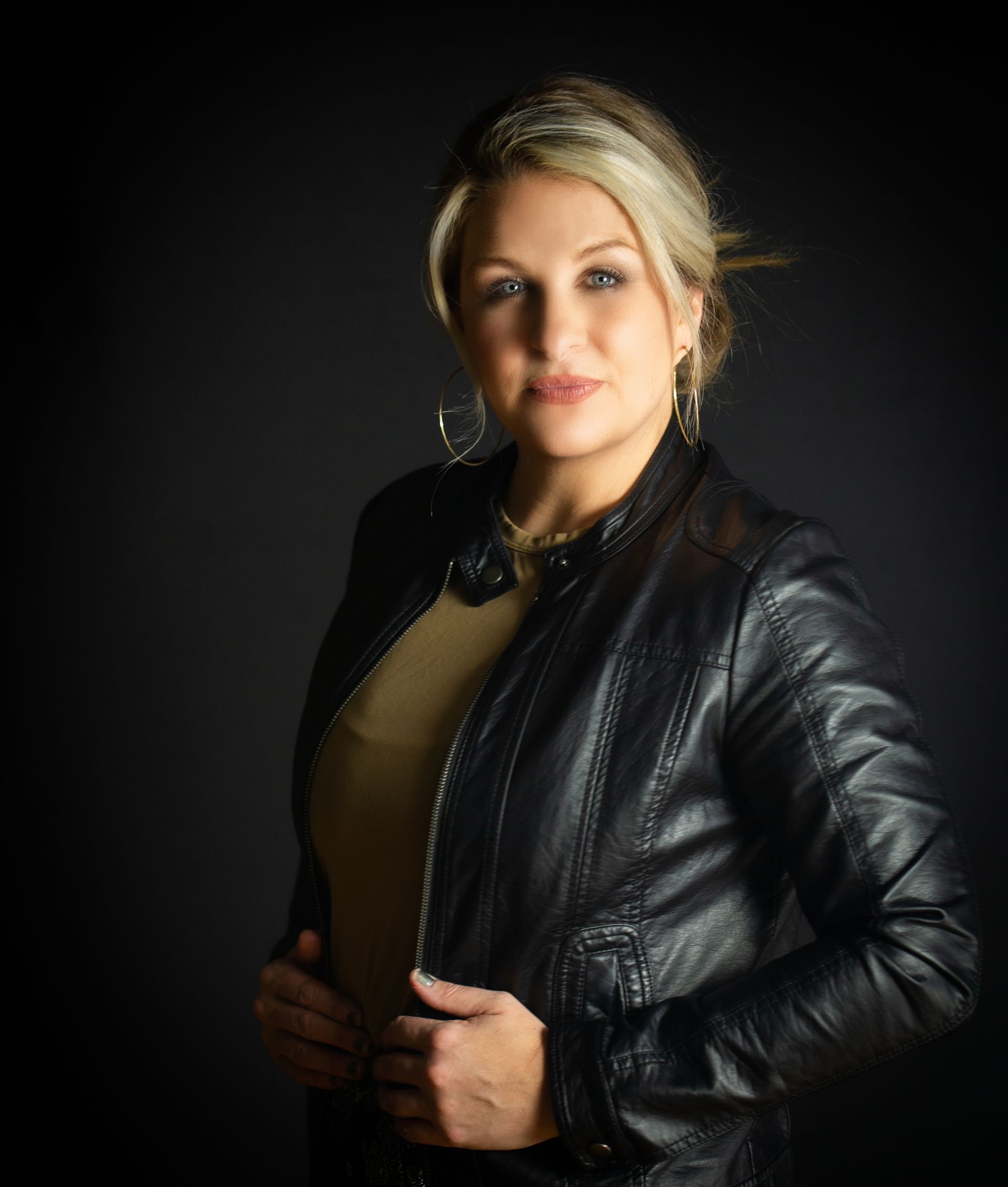 Blonde woman in leather jacket, tan shirt. Posing against a dark background, looking at the camera.