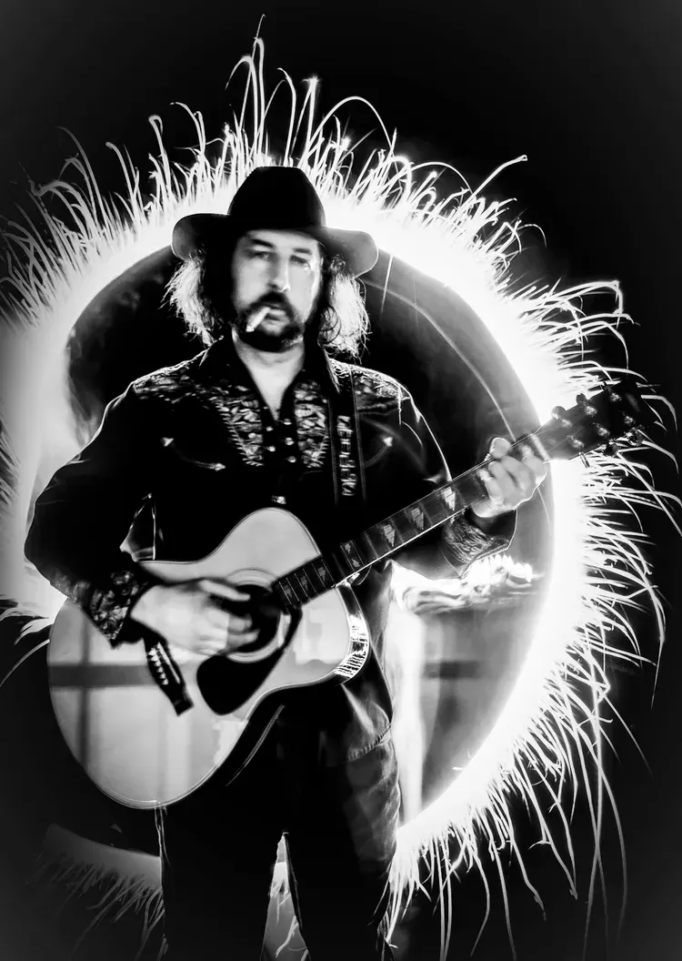 Man playing guitar with a light circle effect behind him. He wears a hat and is indoors.