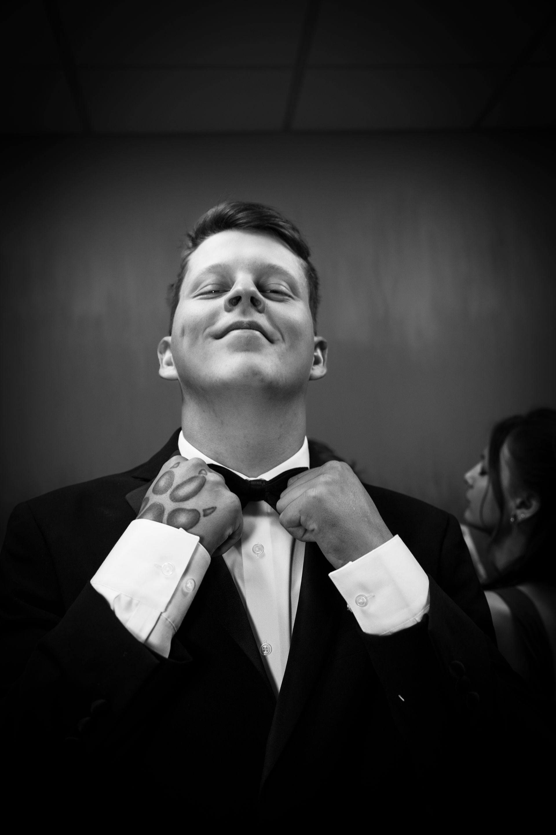 Man in tuxedo adjusting bow tie, smiling, indoors.