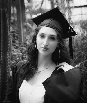 Graduation portrait in black and white: Young woman in cap and gown, smiling, posed in a lush, green environment.