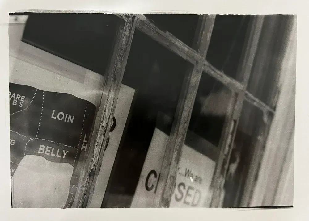 Black and white shot of a butcher shop window. Visible signs include a meat cut chart and a 