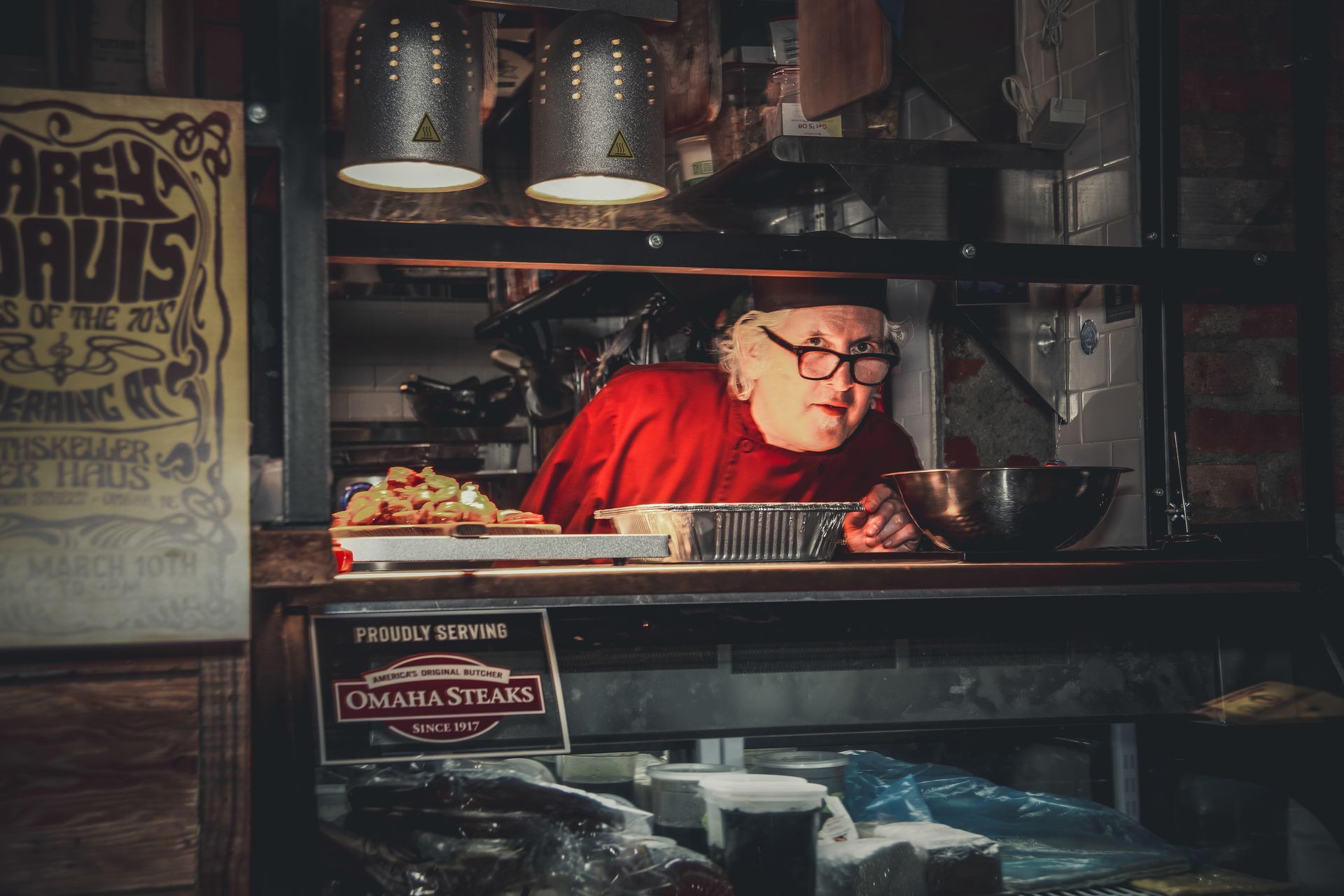 Man in red shirt leans over counter, preparing food in a dimly lit restaurant.