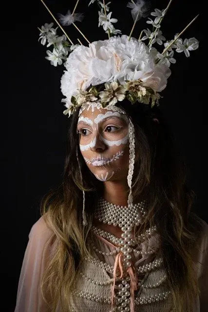 Woman with white face paint, floral crown, and pearl necklace, against a black backdrop.