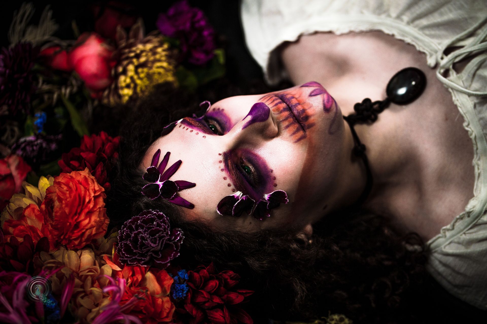 Woman with Dia de los Muertos makeup, surrounded by colorful flowers. Dark background, wearing off-white top.