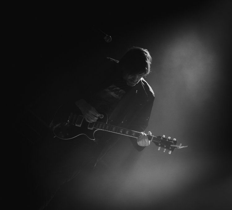 Guitarist playing a black electric guitar, lit by a spotlight, in a dark setting.