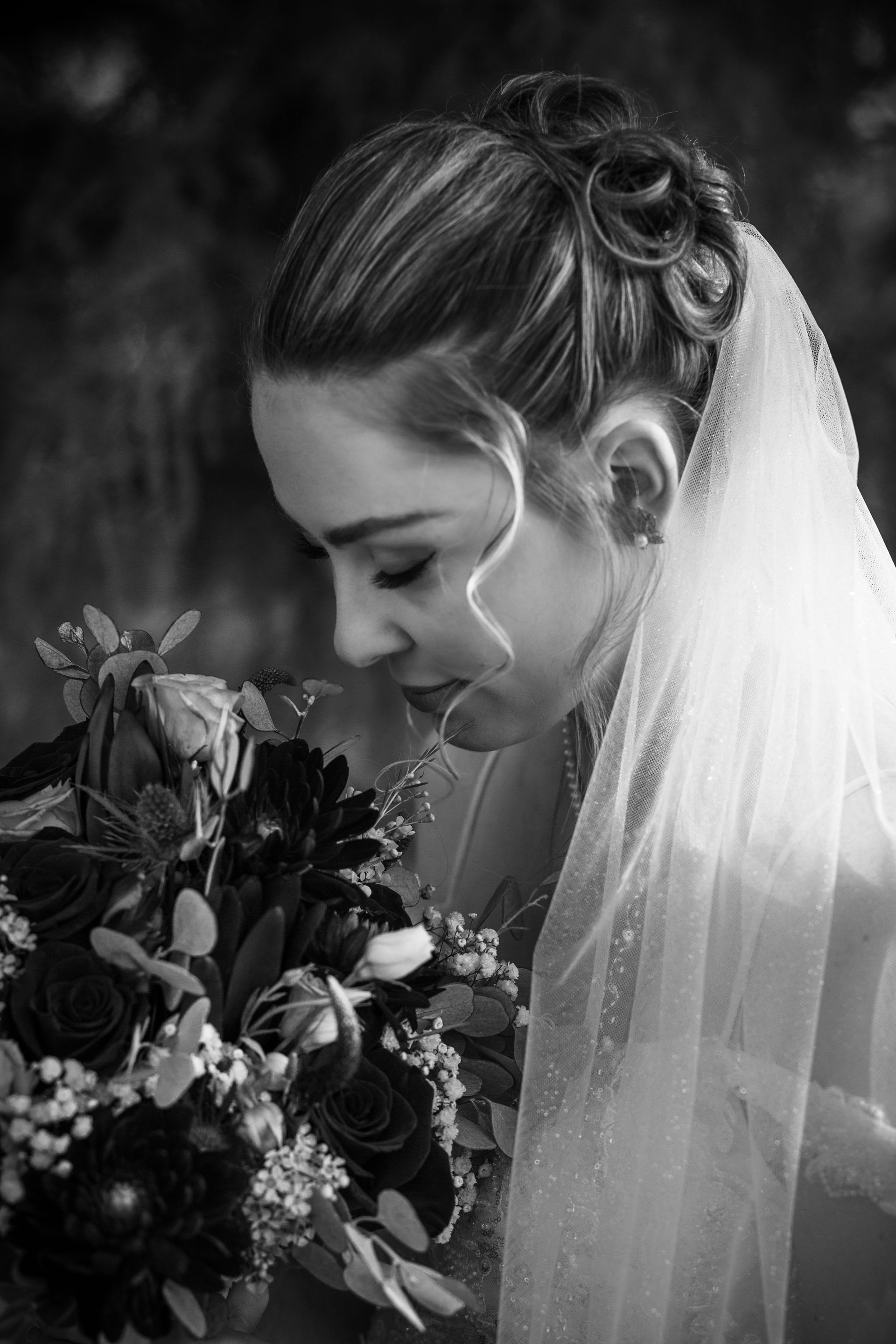 Bride in profile, holding bouquet, looking down. Black and white photo, outdoor setting.