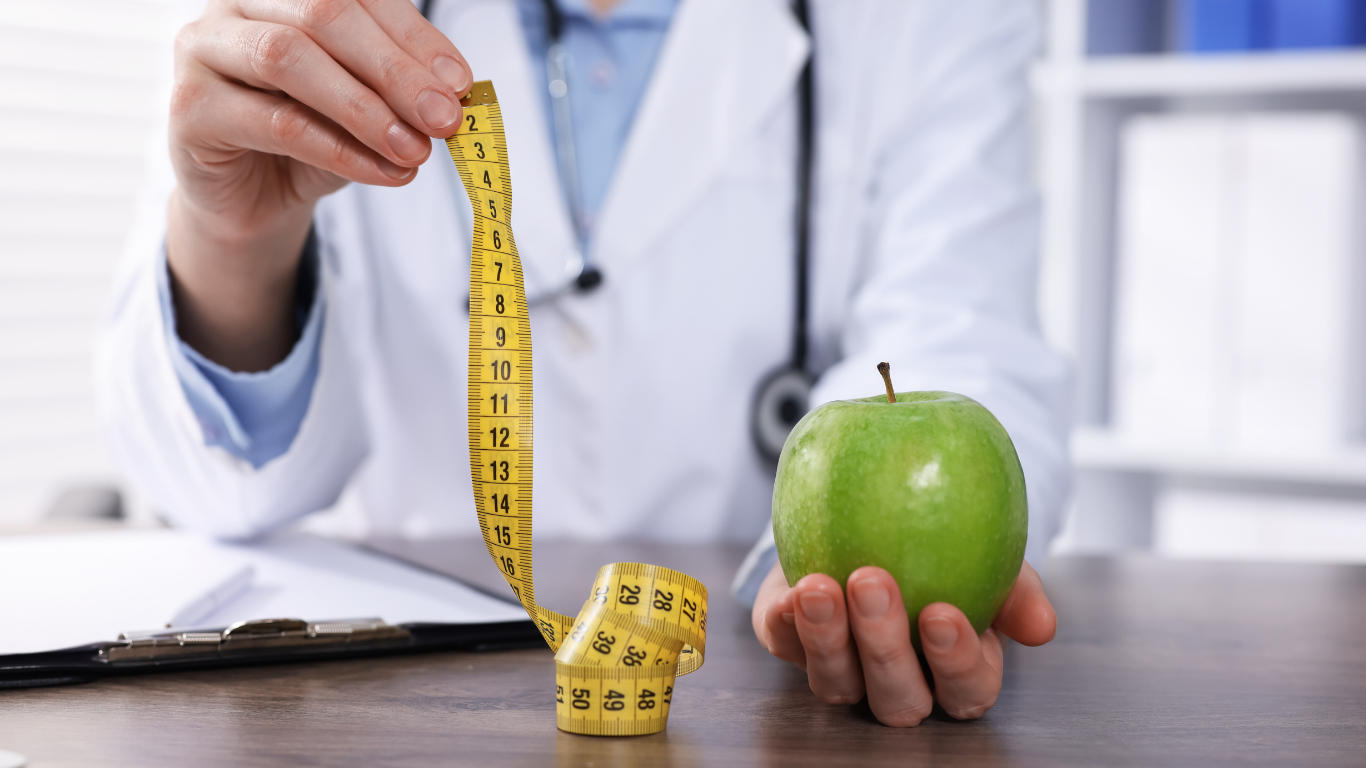 A person in a white lab coat holds a measuring tape and a green apple, sitting at a desk with a medical folder.