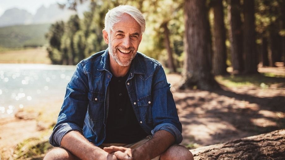 A smiling person wearing a denim shirt sits on a log outdoors near a lake and trees.