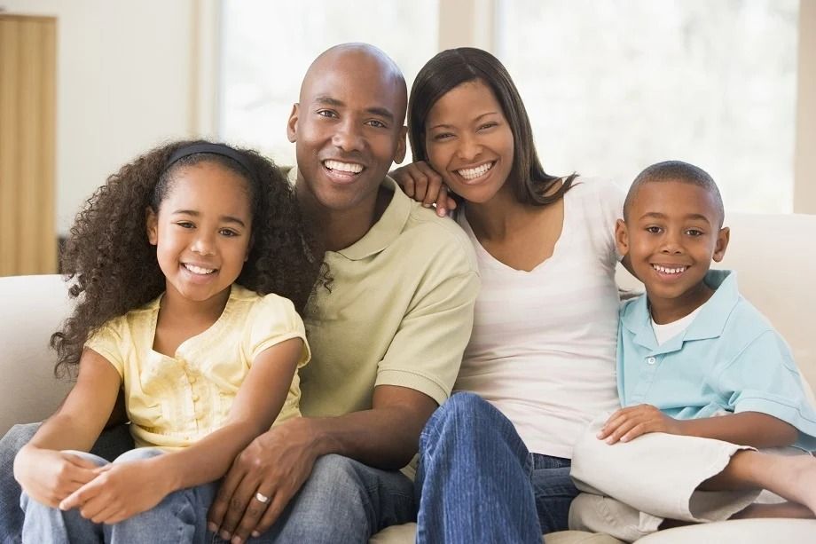 A smiling family of four sitting together on a light-colored couch in a brightly lit room.