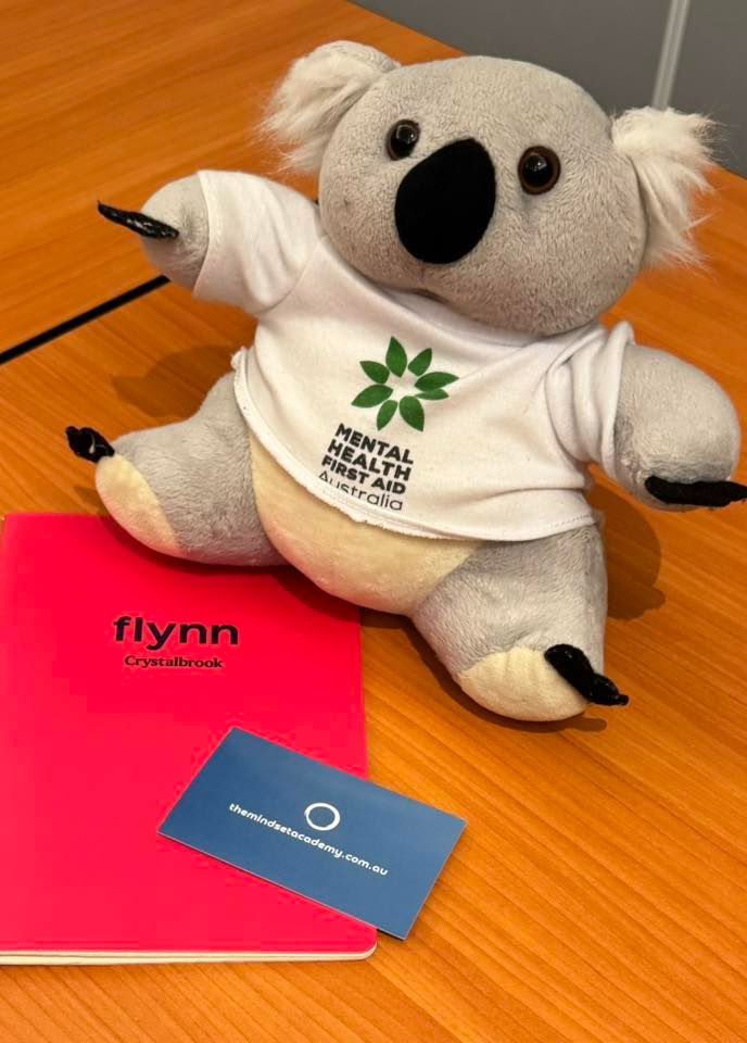 Koala plushie wearing Mental Health First Aid shirt, next to a pink notebook and blue card. Koala plushie wearing Mental Health First Aid shirt, next to a pink notebook and blue card.