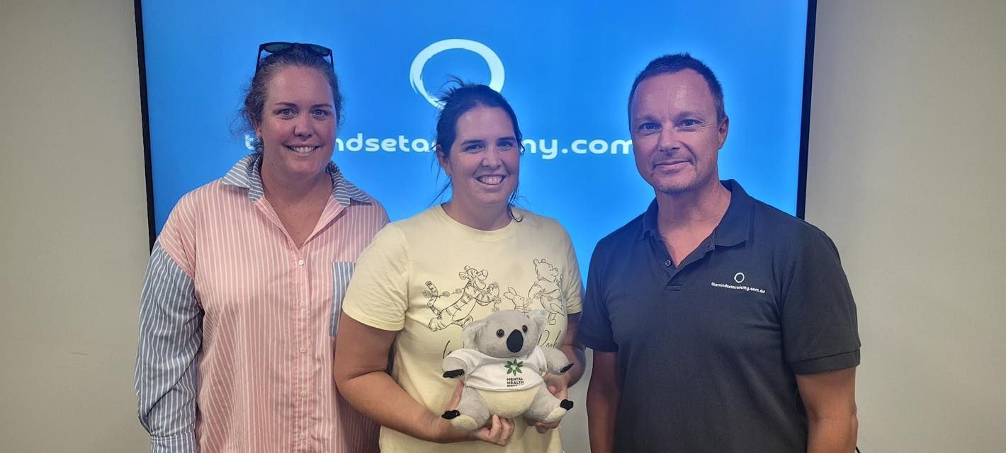 Three people standing in front of a blue screen. A woman holds a stuffed koala, everyone is smiling.
