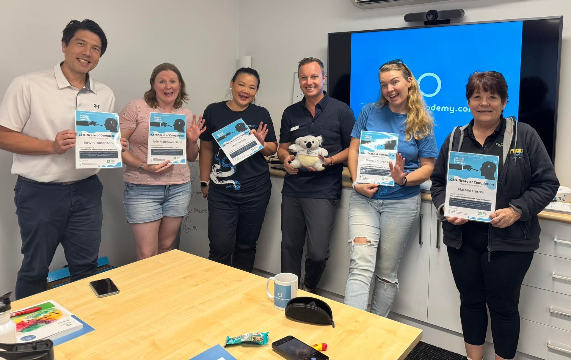 Group of people holding papers in a room. Some smile and show excitement. Blue logo on a screen behind them.