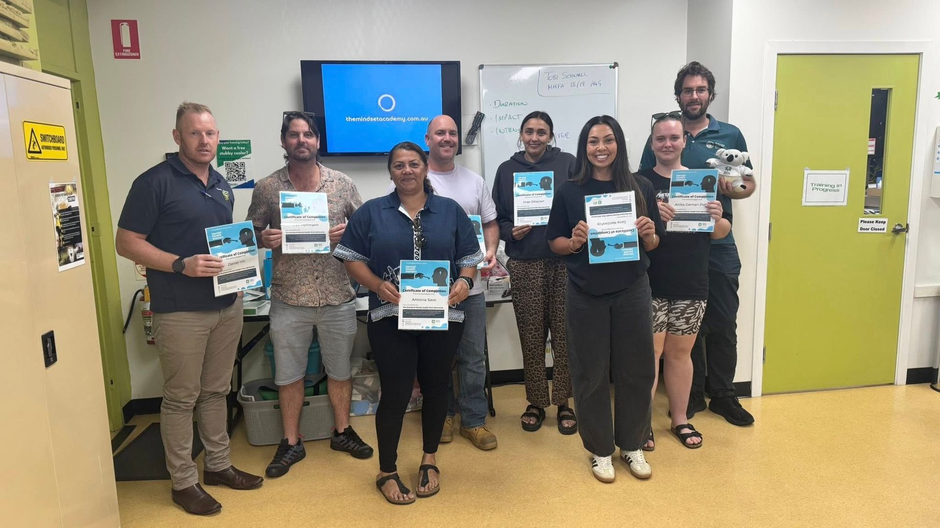 Group of people holding certificates, smiling in a room with a whiteboard and screen.