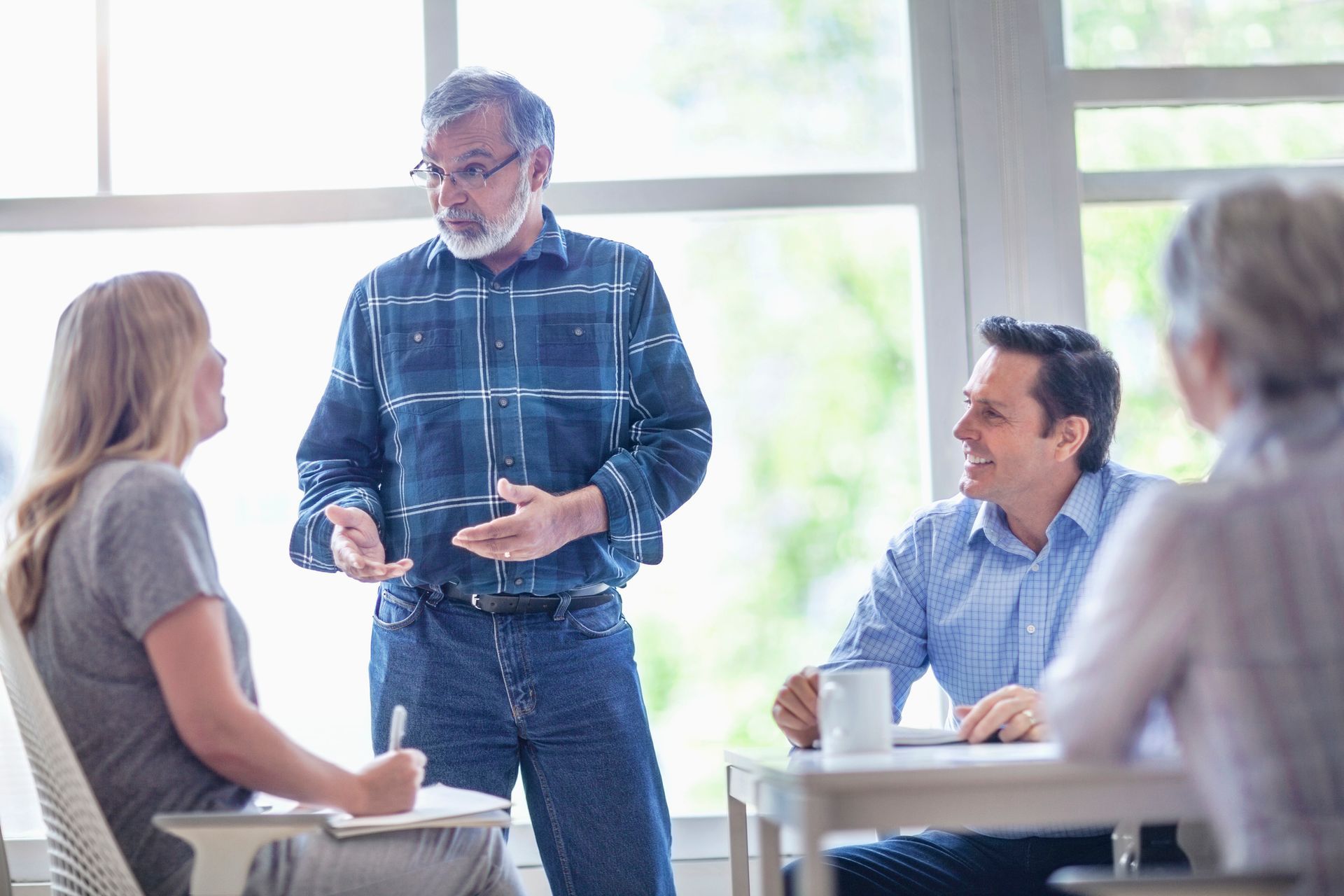 Group of people in a meeting, one man standing and speaking, others seated at a table. Group of people in a meeting, one man standing and speaking, others seated at a table.