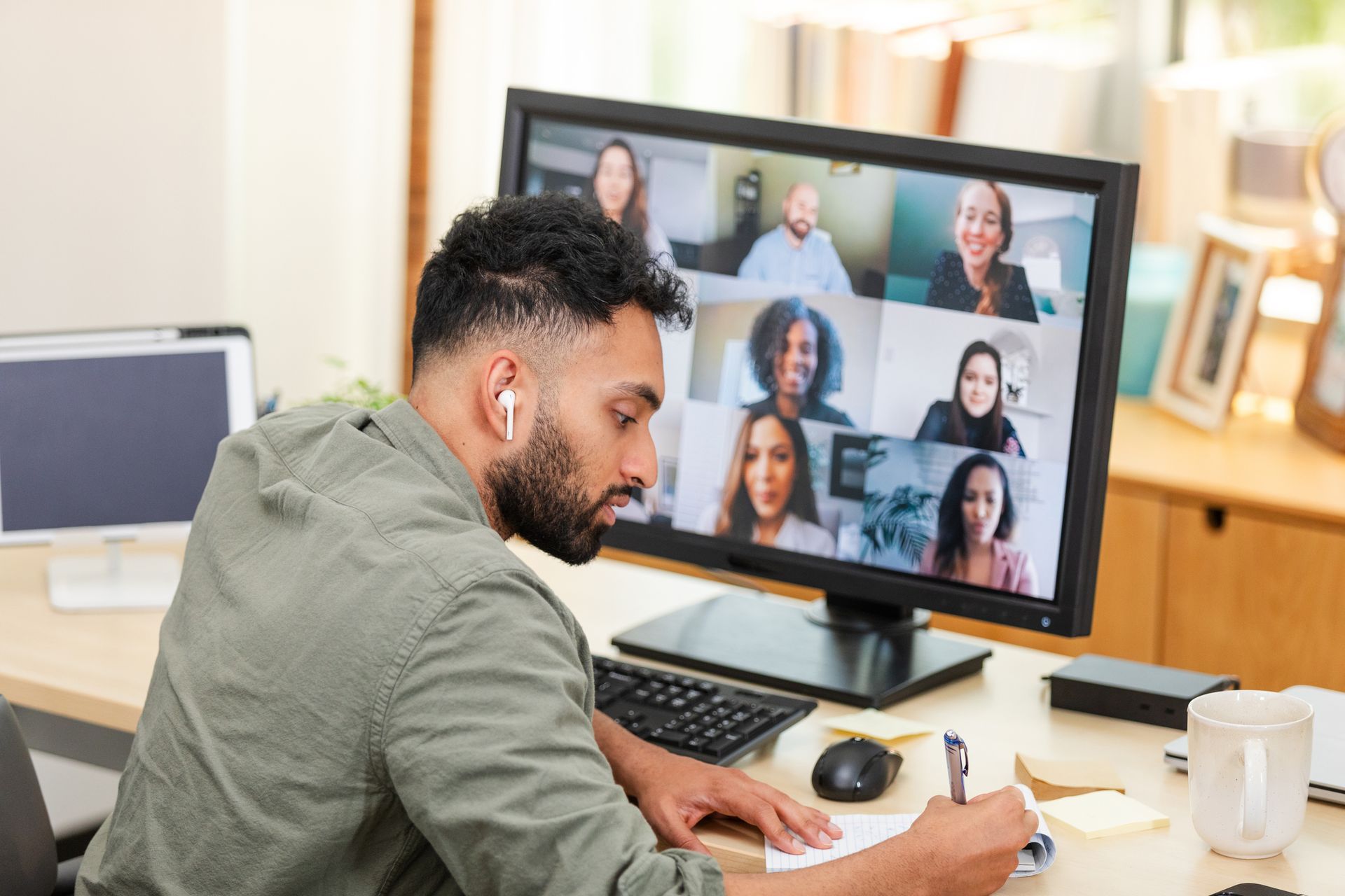 Man in a video conference, writing notes at a desk with a computer displaying other participants. Man in a video conference, writing notes at a desk with a computer displaying other participants.