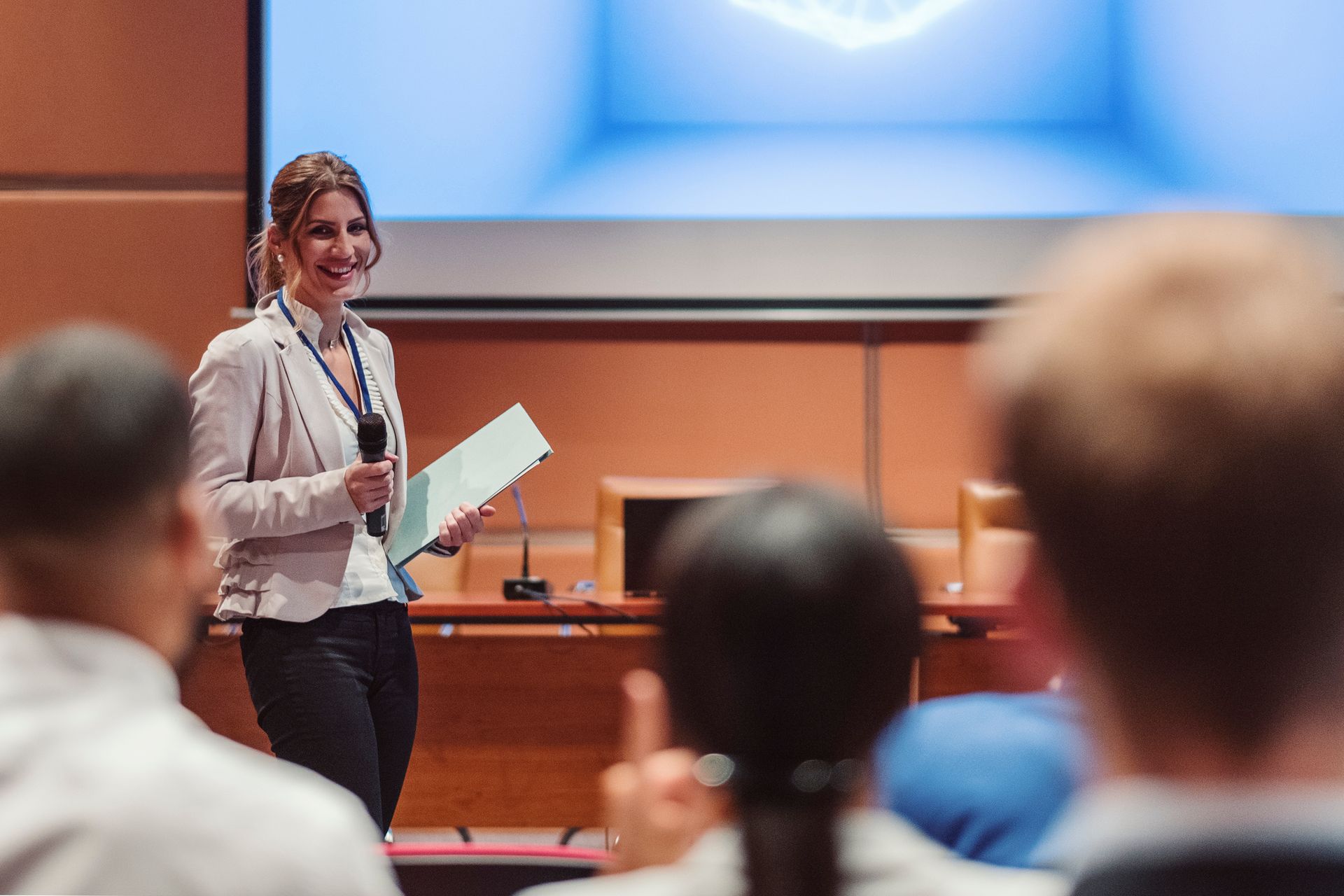 Woman speaking at a conference, holding a microphone and papers, with a screen in the background. Woman speaking at a conference, holding a microphone and papers, with a screen in the background.