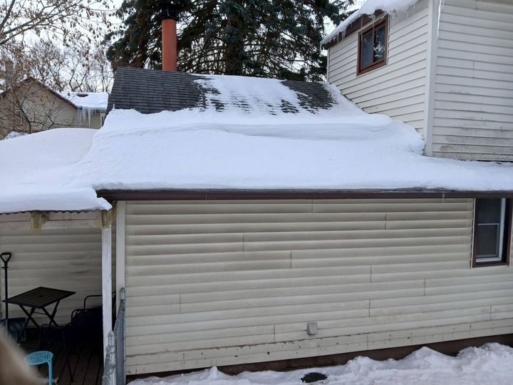 Snow-covered garage roof beside a house, with a chimney and a small patio area underneath.