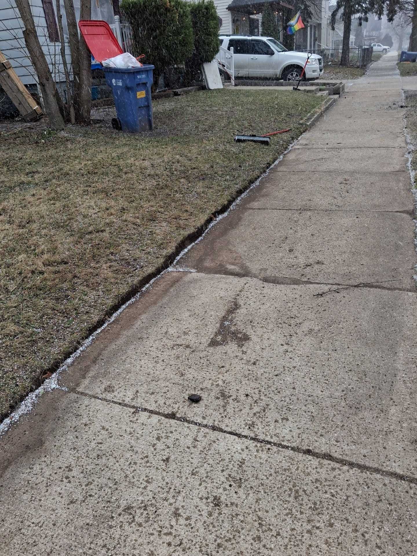 Cracked sidewalk beside a grassy strip with trash bins and parked cars in a neighborhood.