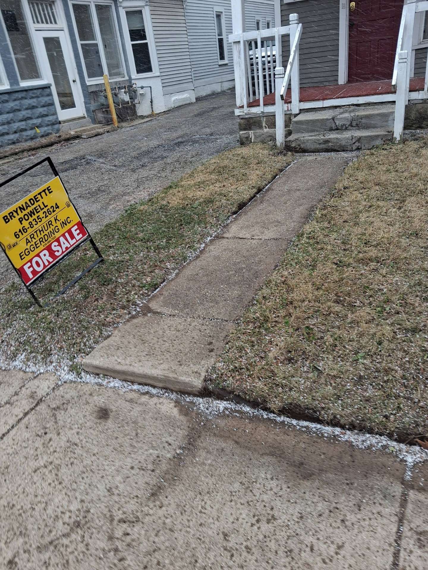 Concrete walkway by houses with a yellow “For Sale” sign in a front yard.