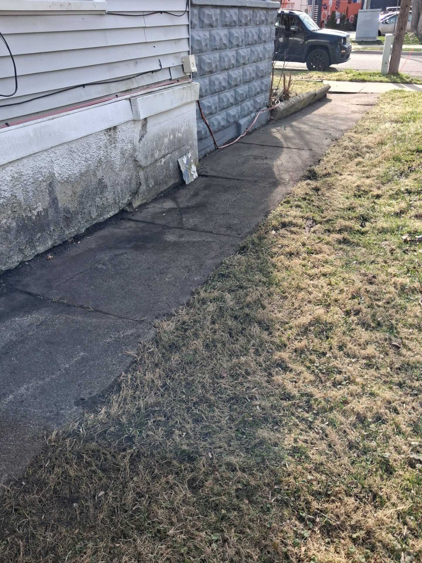 Cracked concrete walkway beside a house, with a grassy yard and a parked vehicle in the background.