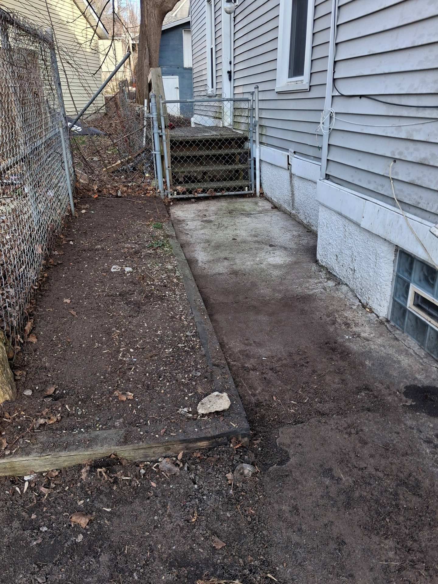 Narrow concrete side path beside a house, leading to back steps and a chain-link fence.