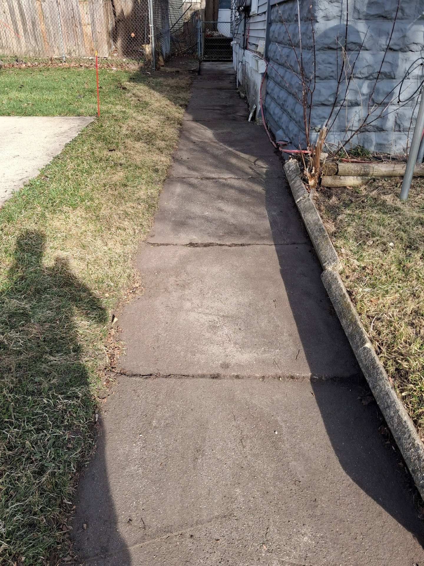 Concrete sidewalk beside a house and lawn, with a narrow walkway leading past a blue wall.