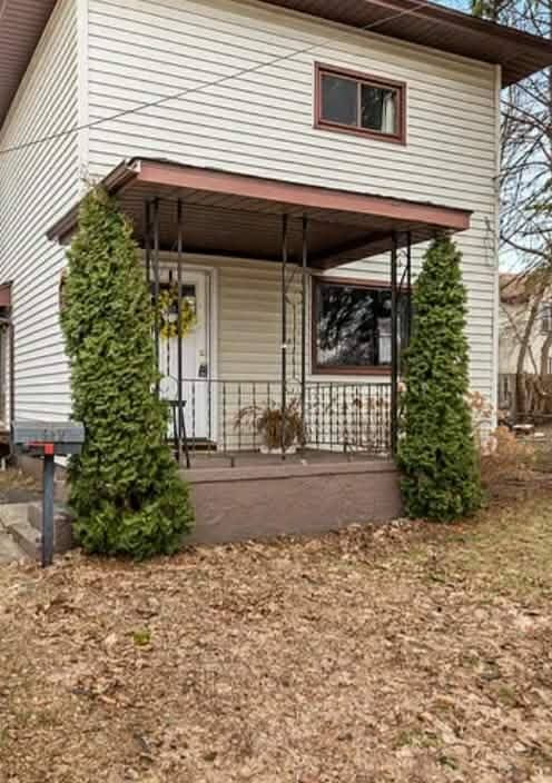Small two-story house with a covered front porch, beige siding, and two tall evergreen shrubs.