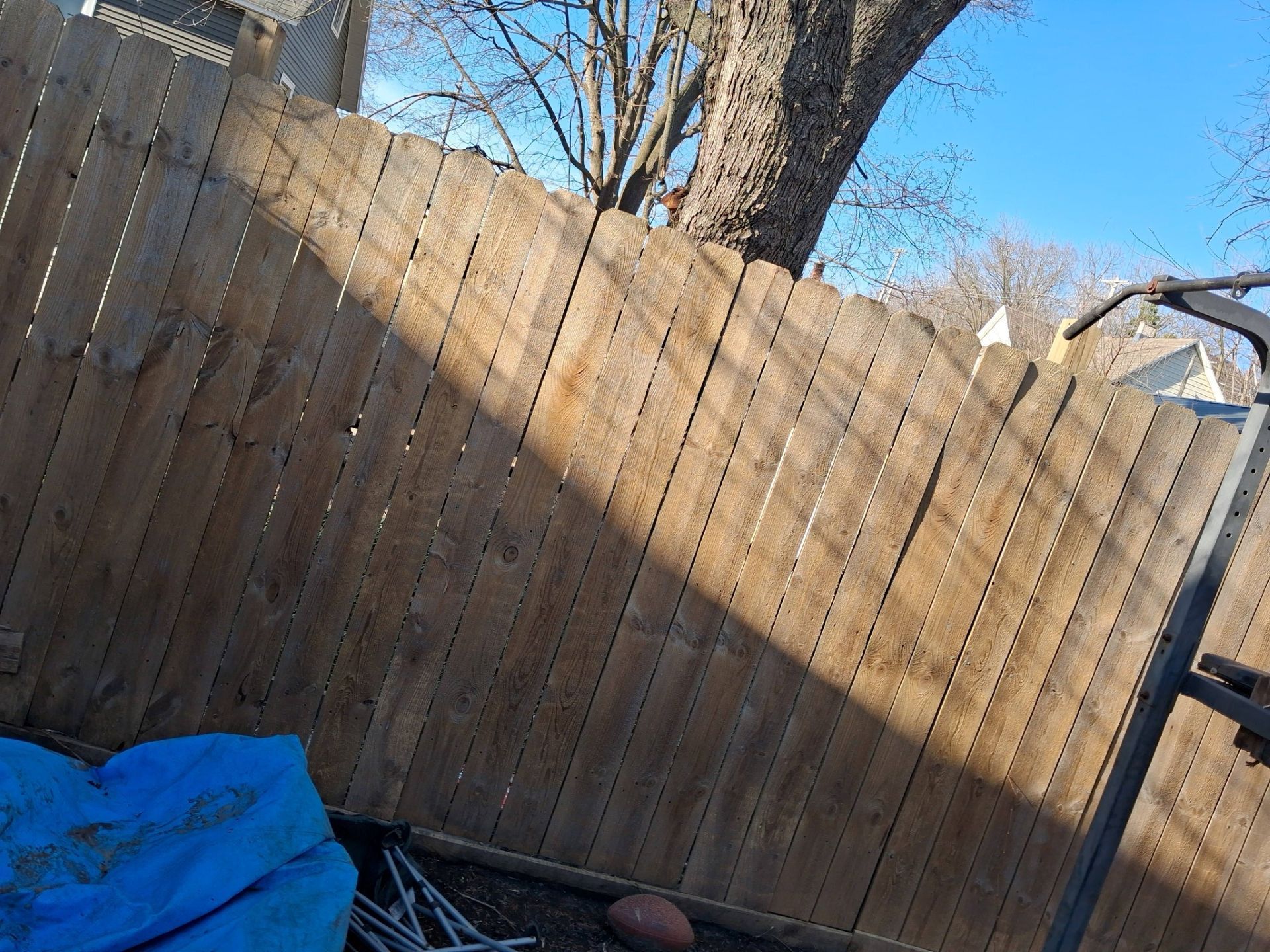 Tilted wooden fence in a backyard, with a blue tarp and a basketball in the foreground under a tree.