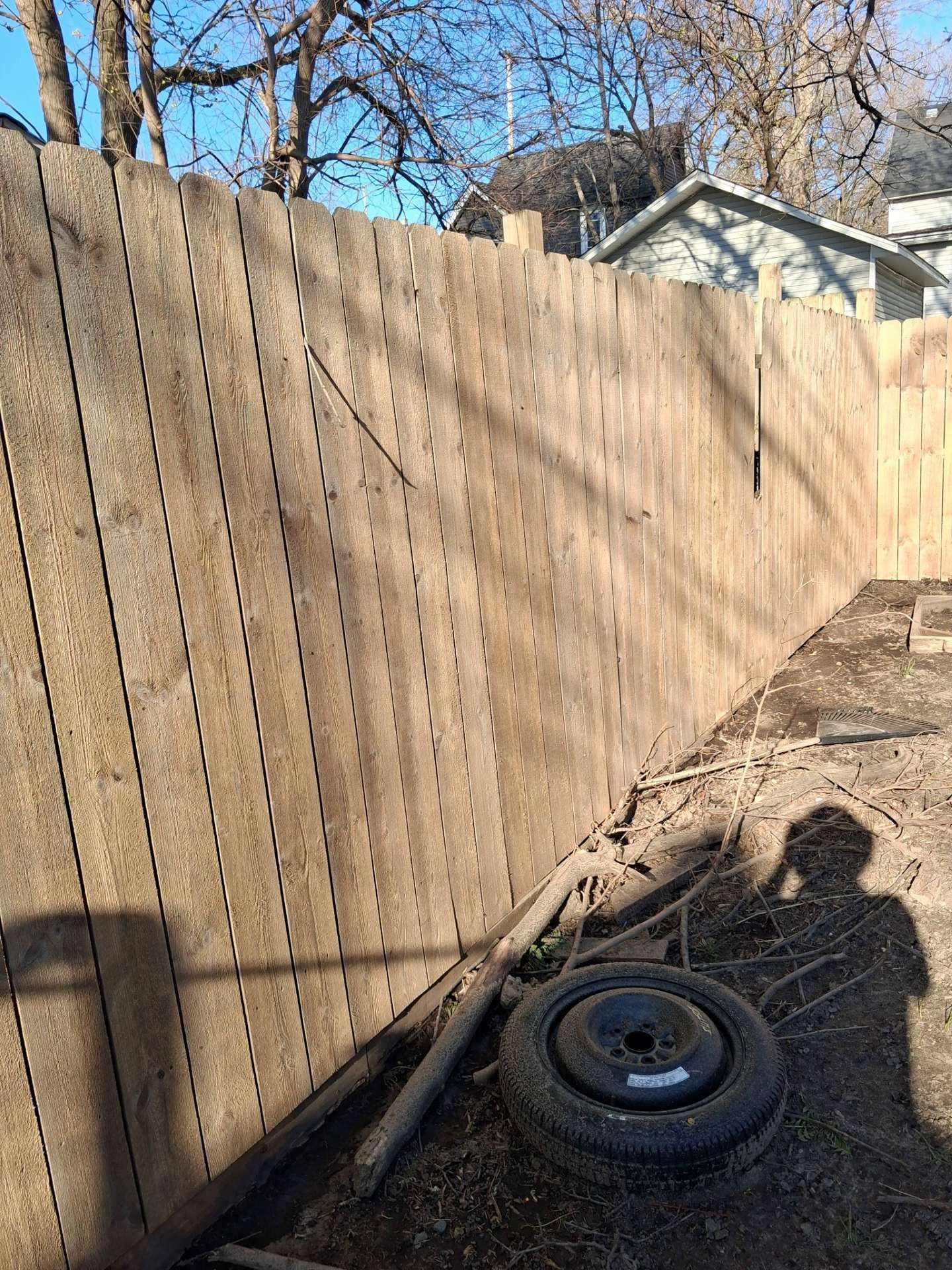 Tall wooden fence beside a dirt path with a spare tire on the ground and trees overhead.