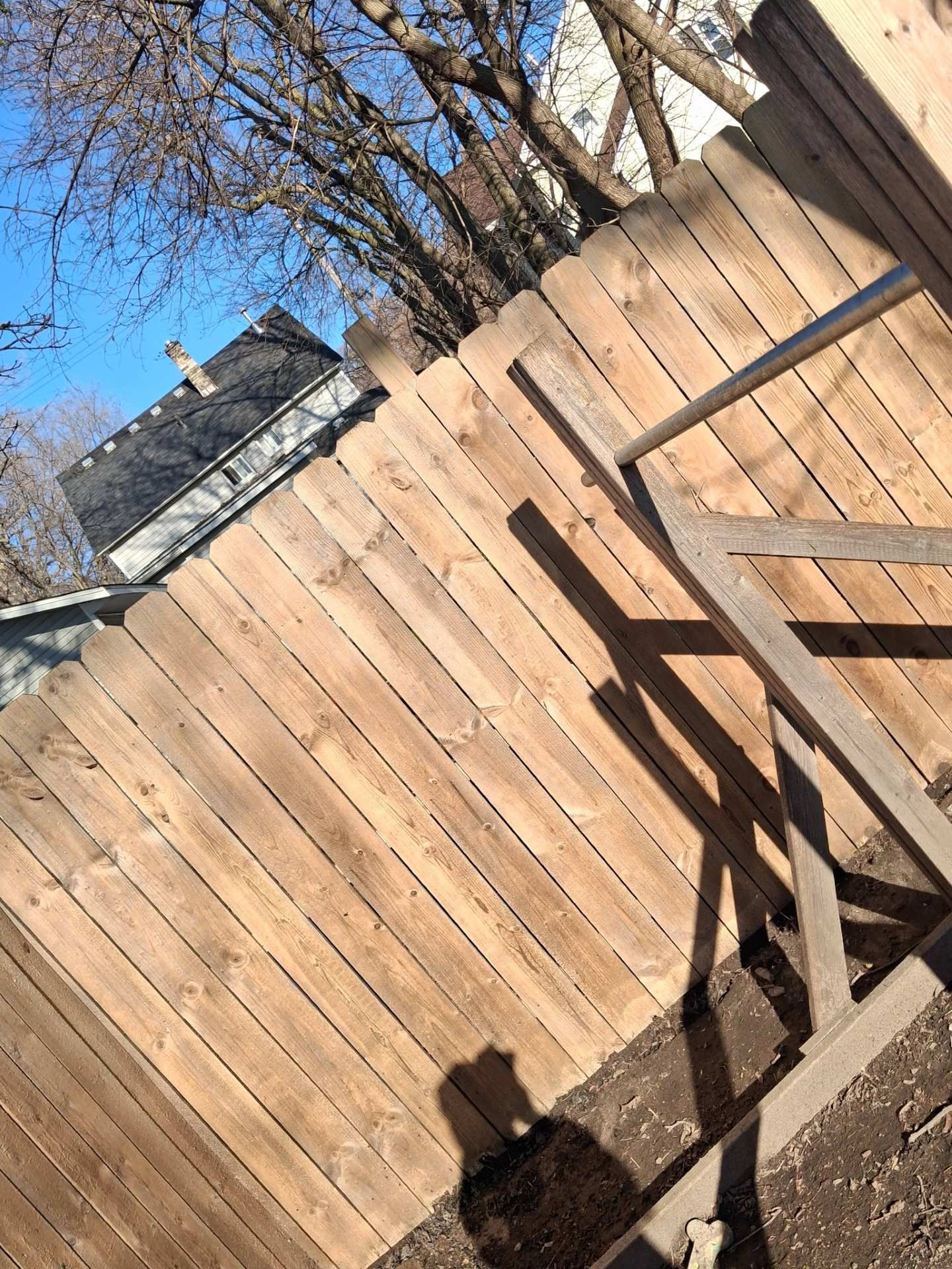 Tilted view of a wooden backyard fence with leafless trees and blue sky behind it, with a shadowed deck railing in front