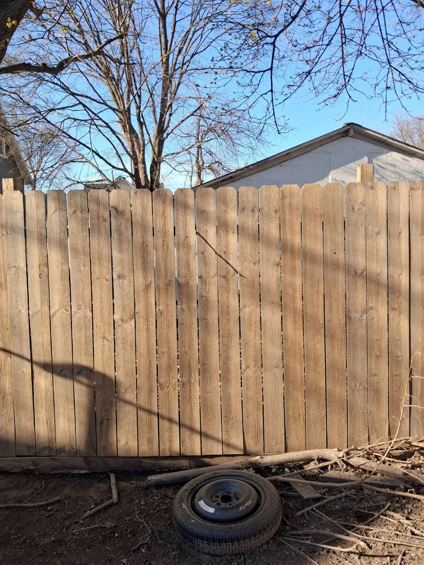 Wooden privacy fence in a backyard with bare trees, a house roof, and a tire on the ground.