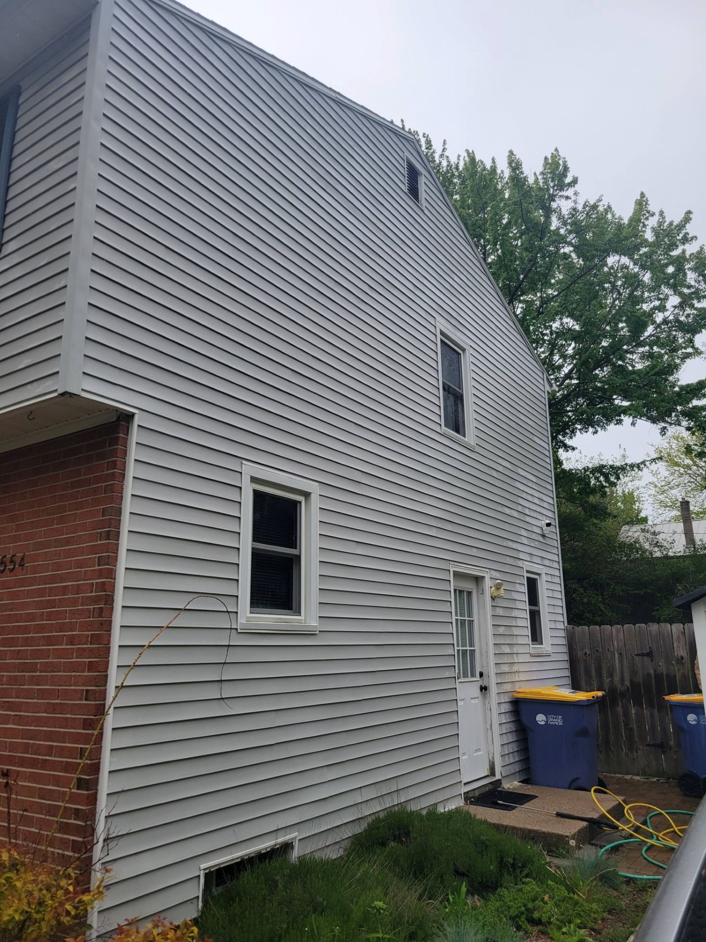 Side view of a gray-sided house with a red brick corner, windows, and a blue recycling bin near a fence.