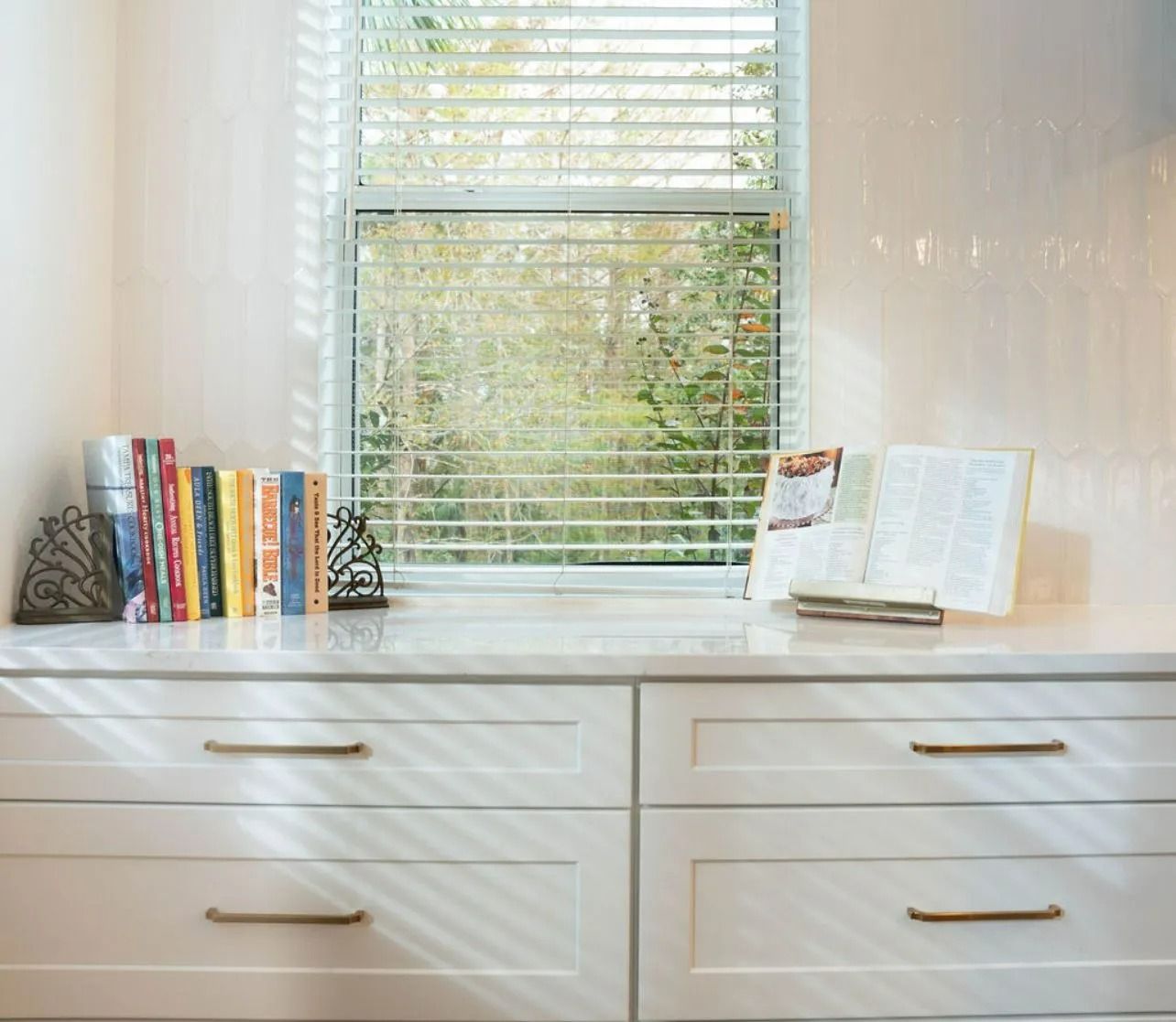 A window with blinds is behind a dresser with books on it.