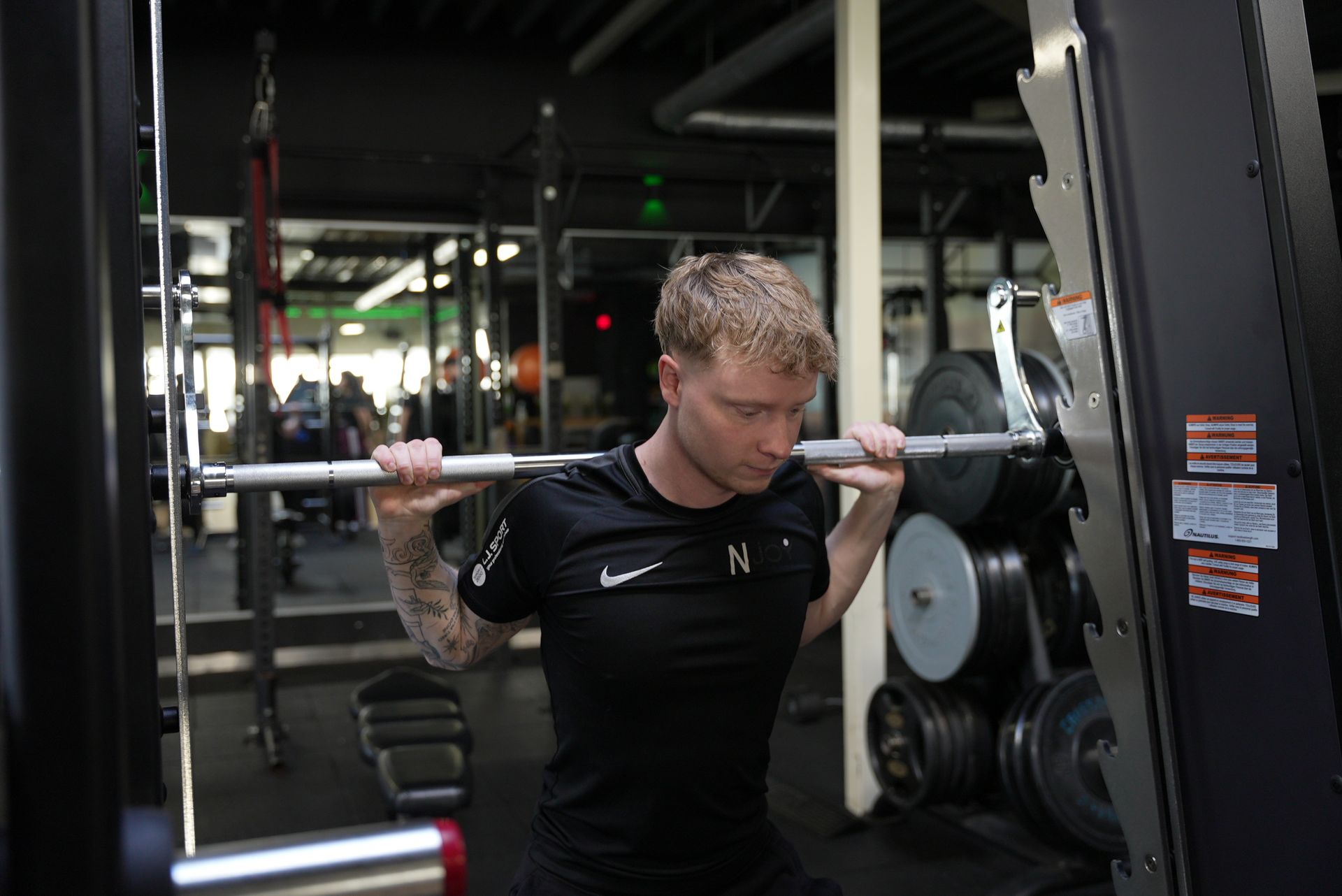 Een man gebruikt fitnessapparatuur, lachend, terwijl een trainer toekijkt. Sportschoolomgeving.