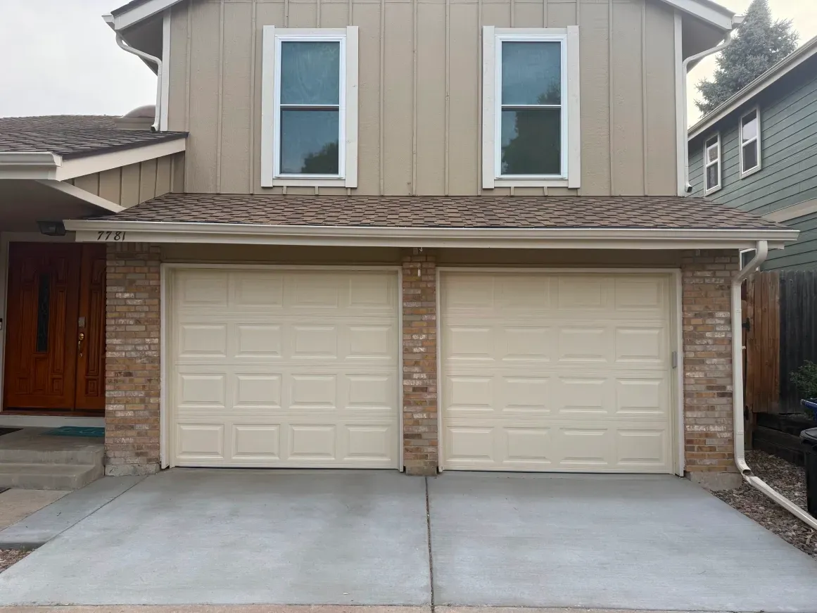 A tan two-story house with two beige garage doors, brick accents, and a wooden front door.