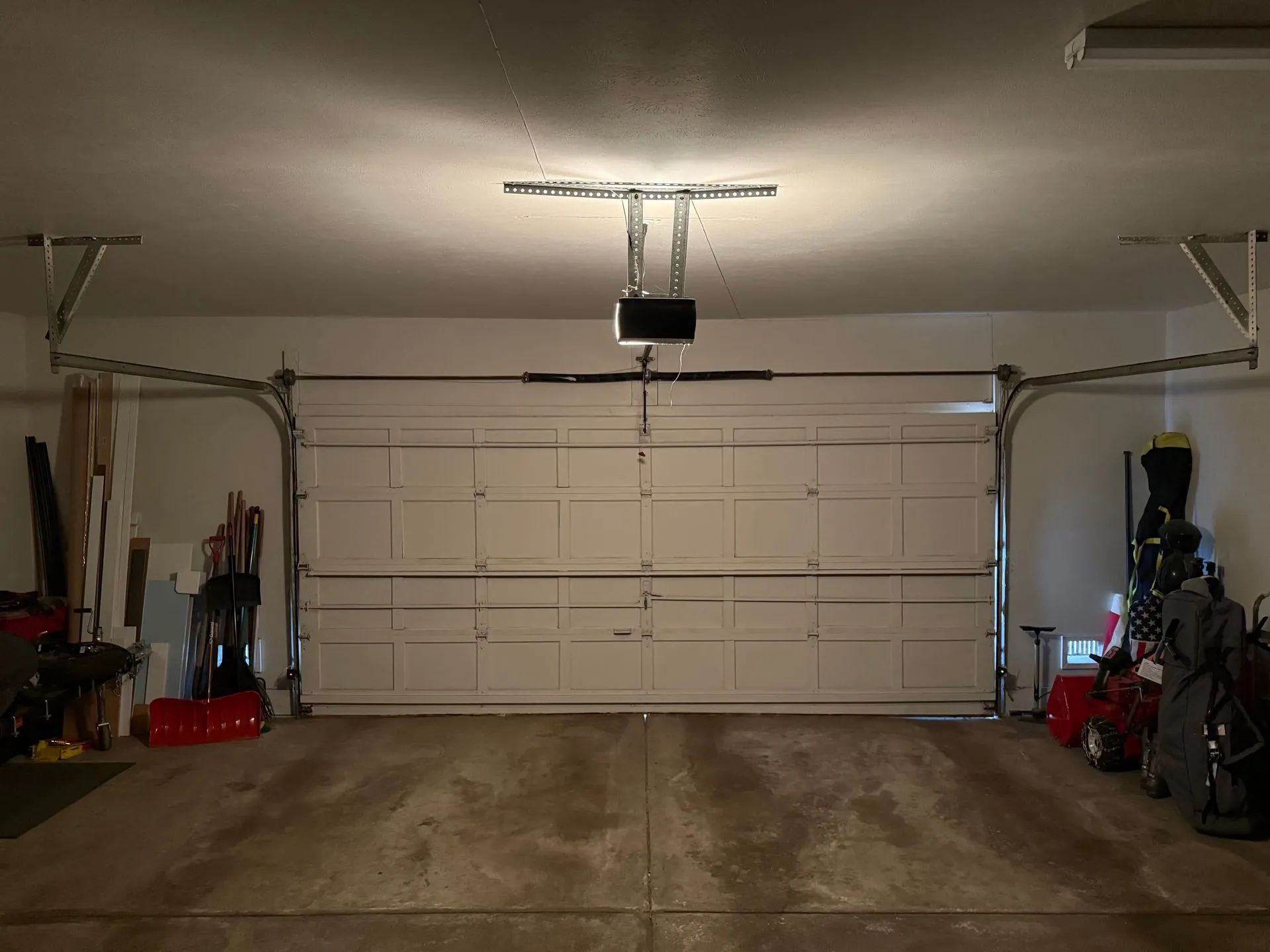 Empty two-car garage with closed white door, ceiling lights, and tools/storage along both sides.