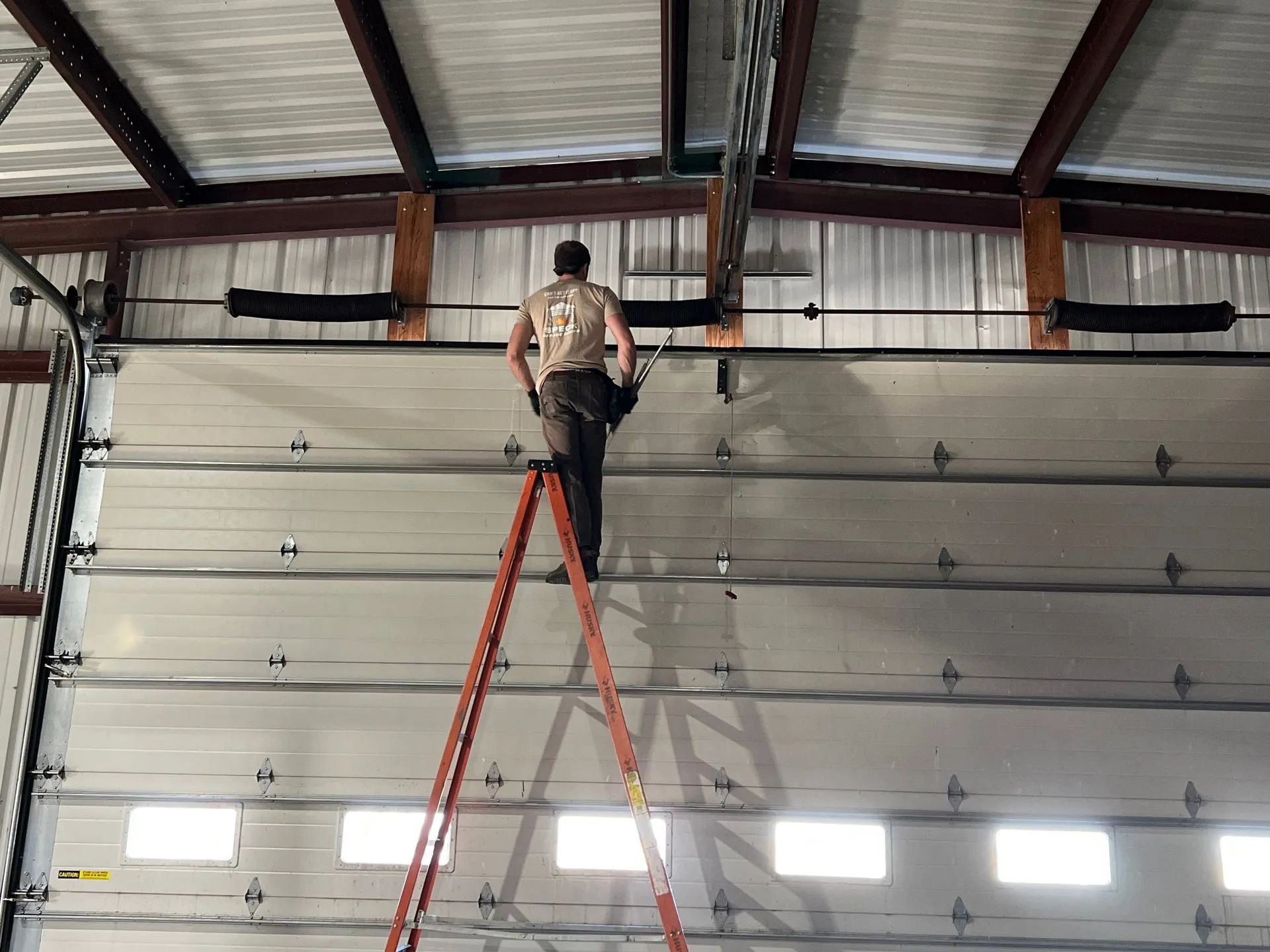 Person standing on a ladder working on a large garage door inside an industrial building.
