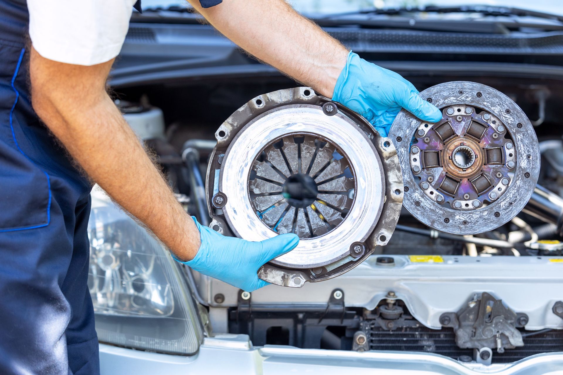 An auto technician is holding a used car pressure plate & clutch disc in front of a car engine.