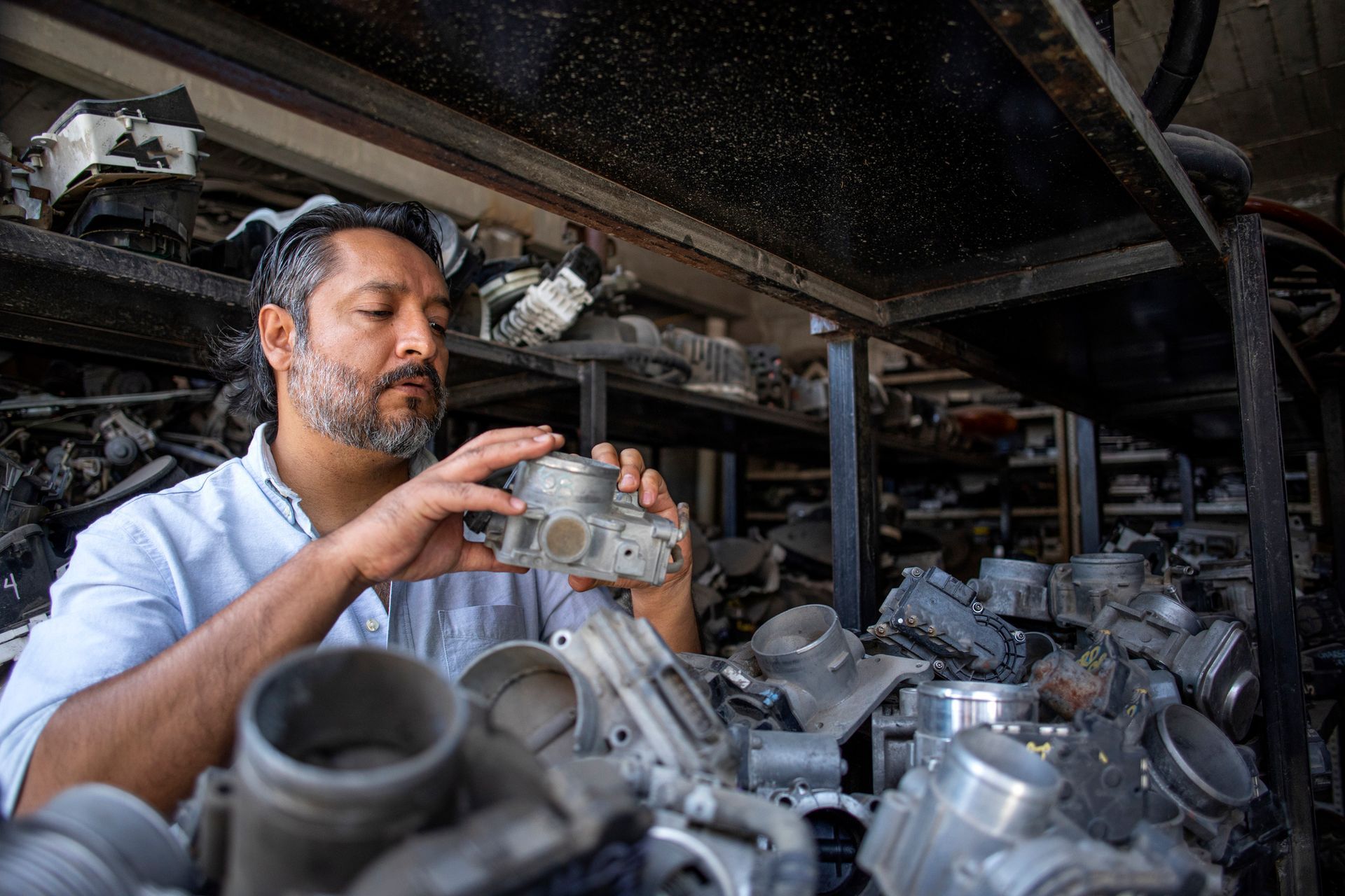 Man inspects an auto part as he sits in front of a table full of parts, inside a parts shop.